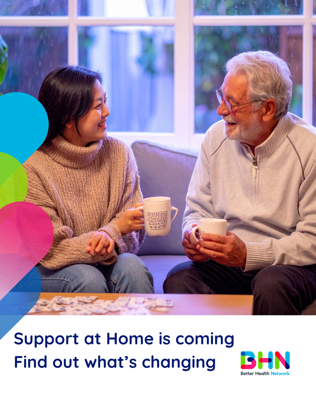 Smiling woman and older man sitting on couch holding coffee mugs and talking. Puzzle pieces spread out on coffee table.
